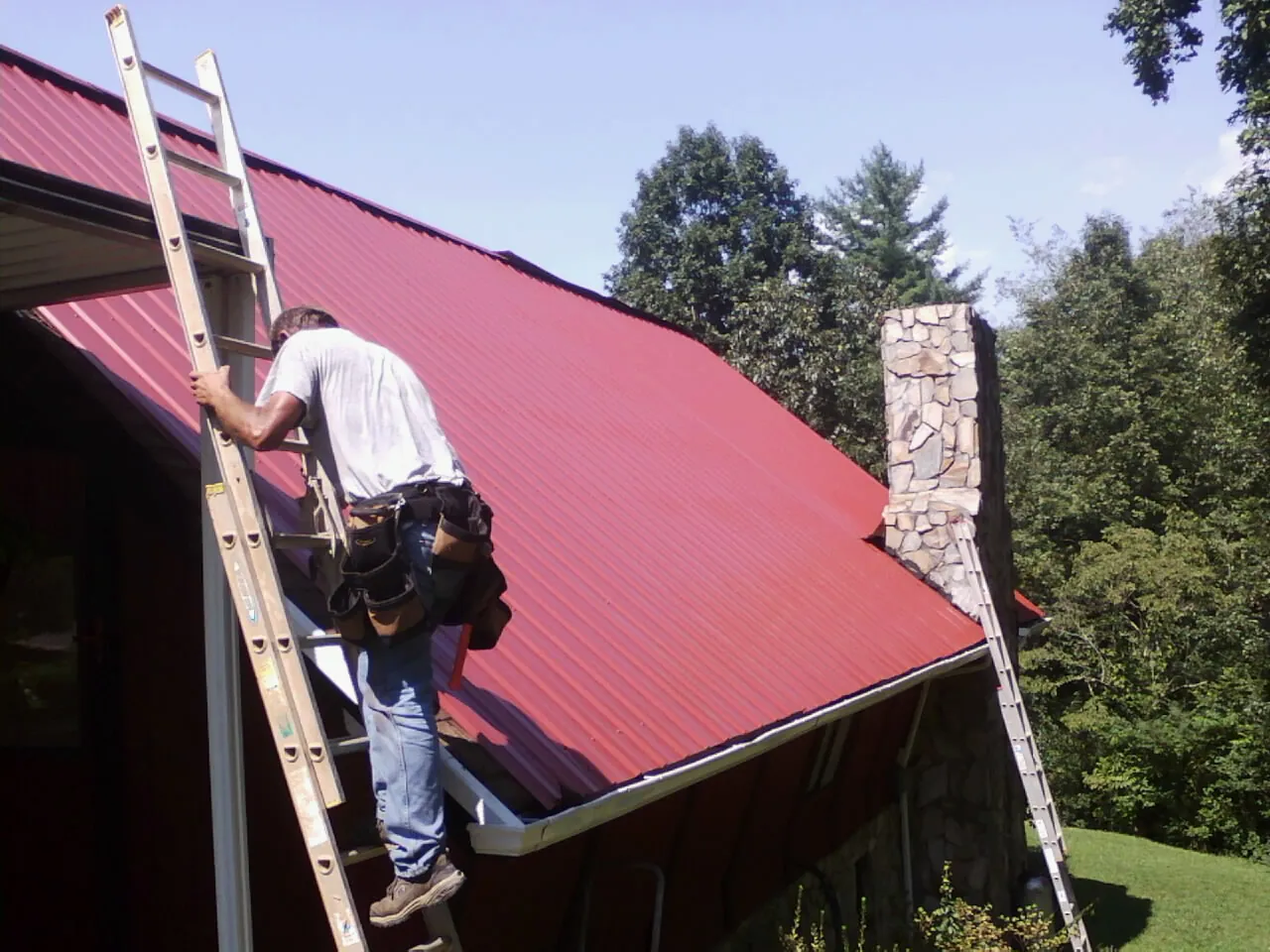 Roofer on a ladder beside a red metal roof and stone chimney — Soffit & Fascia Repair by Roofing Brothers in Viola